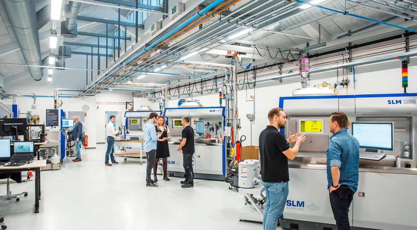 Mænd og kvinder foran metal 3D-printere i produktionshal